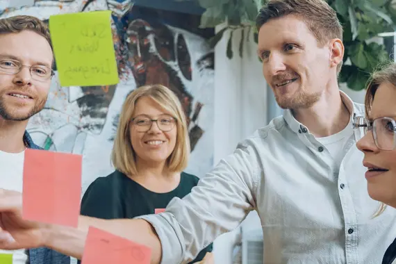Vier Personen in einem Büro, die gemeinsam bunte Haftnotizen an eine Glaswand kleben und diskutieren