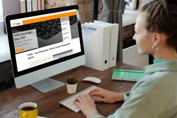 Frau sitzt an einem Schreibtisch und arbeitet an einem Computer mit der TYPO3-Website, die Informationen zu TYPO3 Developer Days zeigt